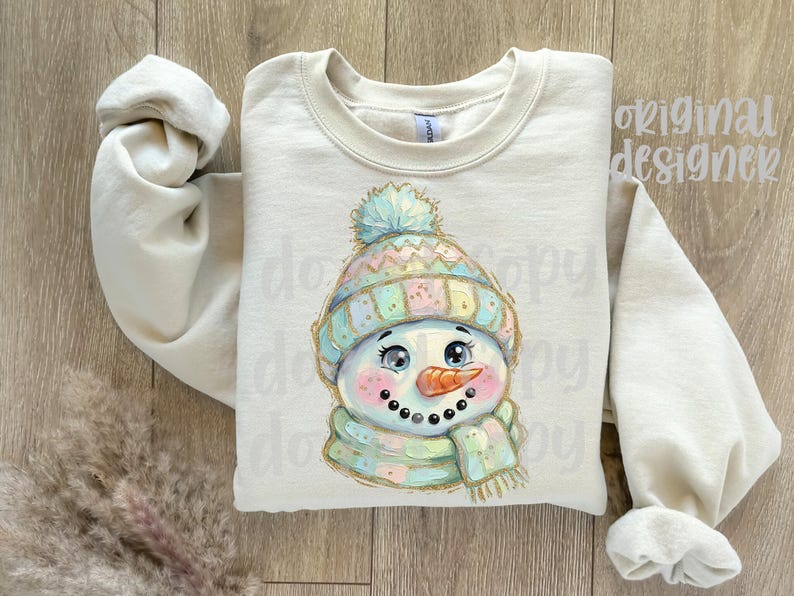 May include: A cream-colored crewneck sweatshirt featuring a central graphic of a snowman wearing a pastel-colored knit hat with a pom-pom and a matching scarf. The snowman has a bright orange carrot nose, rosy cheeks, and large dark eyes.