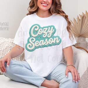 May include: White t-shirt with the text "Cozy Season" in a teal and cream-colored, textured design. The shirt is worn by a person sitting on a couch. The words "Original Designer" are also visible.