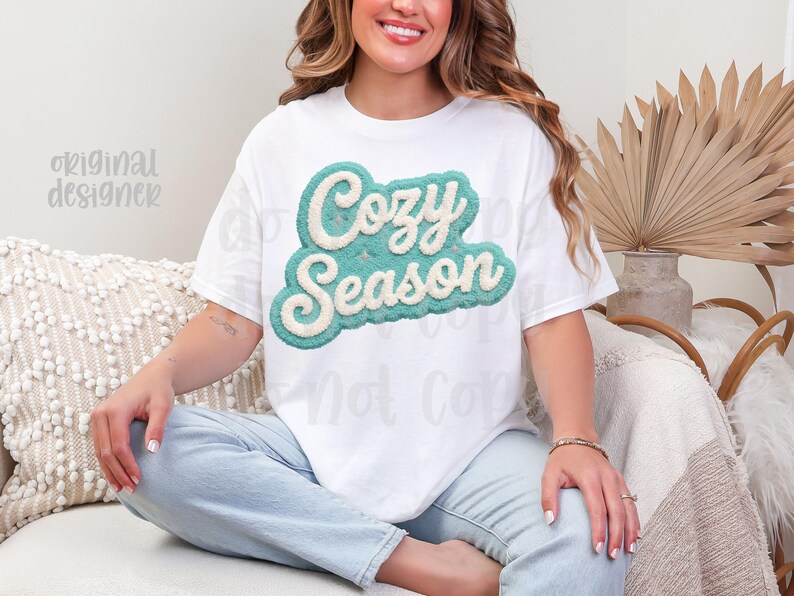 May include: White t-shirt with the text "Cozy Season" in a teal and cream-colored, textured design. The shirt is worn by a person sitting on a couch. The words "Original Designer" are also visible.