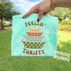 Pu&ograve; includere: T-shirt verde acqua chiaro con le parole "FEELIN' THRIFTY" in carattere retr&ograve; marrone. La maglietta presenta una pila di pirofile vintage gialle, marroni e verde acqua, con accenti di margherite. La scritta "original designer" &egrave; in basso a destra.