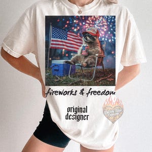 May include: Cream-colored t-shirt featuring a graphic of a raccoon wearing a hat that says "America", holding a can, and sitting in a chair in front of an American flag and fireworks. The text "fireworks & freedom" and "original designer" are also printed on the shirt.