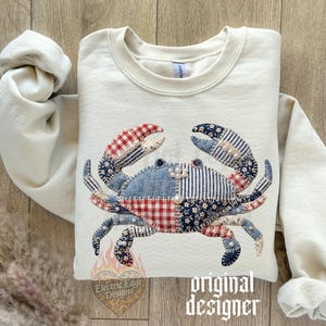 May include: Cream-colored sweatshirt featuring a patchwork crab design. The crab is made of various patterned fabrics in red, blue, and white. The sleeves are rolled up, and the text "original designer" is visible.