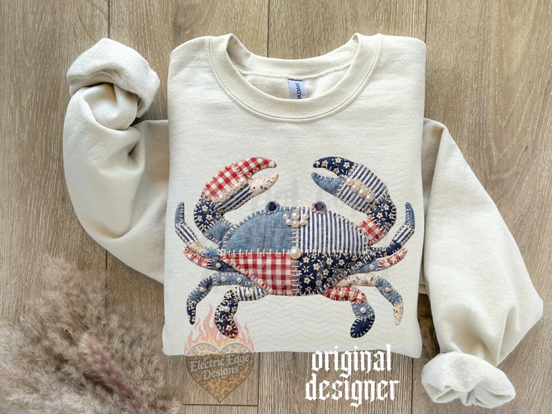 May include: Cream-colored sweatshirt featuring a patchwork crab design. The crab is made of various patterned fabrics in red, blue, and white. The sleeves are rolled up, and the text "original designer" is visible.