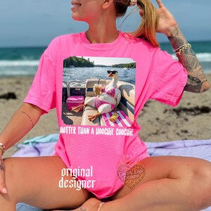 Puede incluir: Camiseta rosa brillante con un gr&aacute;fico de un ganso con gafas de sol y bikini, sosteniendo una bebida y sentado en un bote. El texto "HOTTER THAN A HOOCHIE COOCHIE" est&aacute; debajo. Las palabras "original designer" tambi&eacute;n est&aacute;n impresas.
