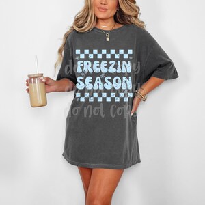 May include: Dark gray oversized t-shirt with the text "FREEZING SEASON" in light blue. The text is set against a checkered pattern with snowflake accents. The model is holding a glass with a straw.
