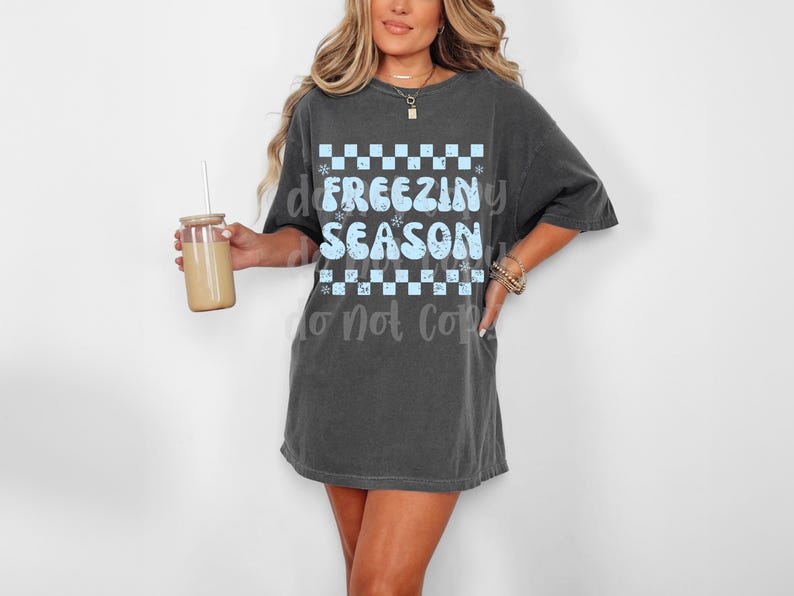 May include: Dark gray oversized t-shirt with the text "FREEZING SEASON" in light blue. The text is set against a checkered pattern with snowflake accents. The model is holding a glass with a straw.