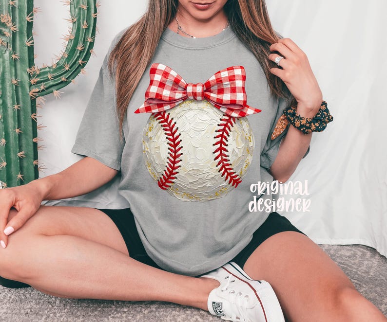 Peut inclure: T-shirt gris avec un motif de baseball et un n&oelig;ud vichy rouge et blanc. La balle de baseball a des coutures rouges et des accents dor&eacute;s. Le texte "original designer" est imprim&eacute; sur le t-shirt.