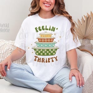 Pu&ograve; includere: T-shirt bianco con un design retr&ograve; con piatti da forno impilati in verde, giallo e turchese. La maglietta ha le parole "FEELIN' THRIFTY" in lettere verdi e marroni. Il design ha un'estetica floreale vintage.