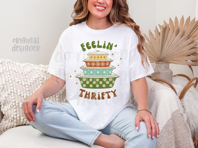 Pu&ograve; includere: T-shirt bianco con un design retr&ograve; con piatti da forno impilati in verde, giallo e turchese. La maglietta ha le parole "FEELIN' THRIFTY" in lettere verdi e marroni. Il design ha un'estetica floreale vintage.