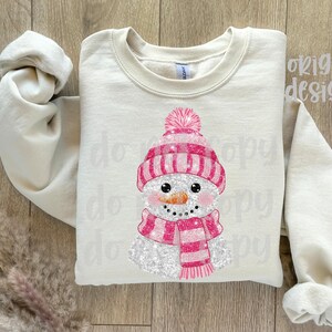 May include: A cream-coloured sweatshirt with a snowman design. The snowman has a pink hat, scarf, and a carrot nose. The design is glittery and festive, suitable for winter or holiday wear.