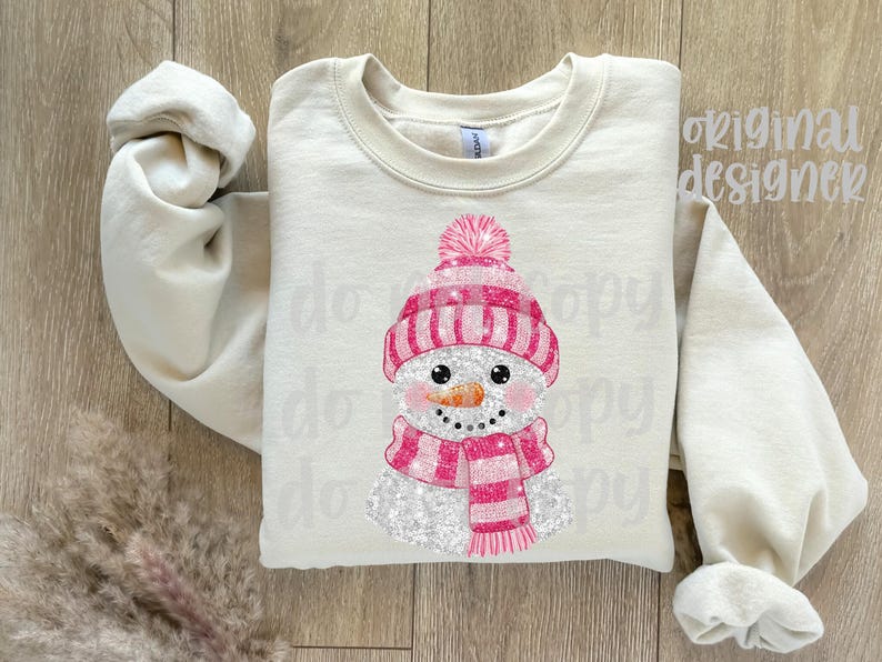 May include: A cream-coloured sweatshirt with a snowman design. The snowman has a pink hat, scarf, and a carrot nose. The design is glittery and festive, suitable for winter or holiday wear.
