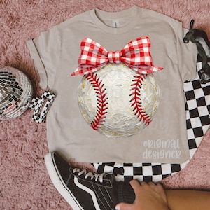 Peut inclure: T-shirt beige avec un motif de baseball orn&eacute; d'un n&oelig;ud vichy rouge et blanc. Le baseball pr&eacute;sente des coutures rouges et des d&eacute;tails dor&eacute;s. Le texte "original designer" est imprim&eacute; sur le t-shirt. Motif &agrave; carreaux noir et blanc visible.