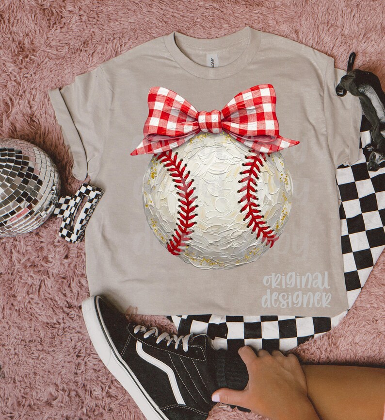 Peut inclure: T-shirt beige avec un motif de baseball orn&eacute; d'un n&oelig;ud vichy rouge et blanc. Le baseball pr&eacute;sente des coutures rouges et des d&eacute;tails dor&eacute;s. Le texte "original designer" est imprim&eacute; sur le t-shirt. Motif &agrave; carreaux noir et blanc visible.