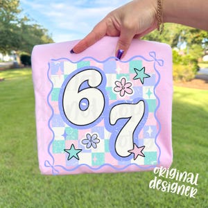 May include: A light pink t-shirt featuring a colorful graphic with the numbers "67" in a retro font. The graphic includes stars, flowers, and a checkered pattern in pastel colors. The words "original designer" are in the bottom right corner.