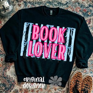 May include: Black sweatshirt with the words "BOOK LOVER" in pink letters, set against a light blue and white striped background. The words "original designer" are printed in white at the bottom. A disco ball and leopard print shoes are in the background.