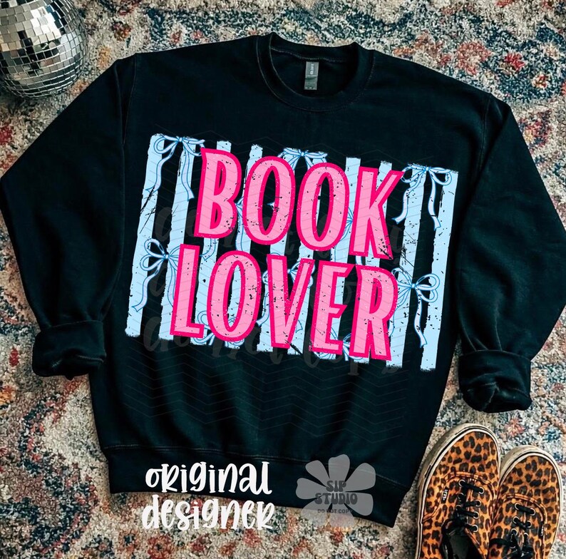 May include: Black sweatshirt with the words "BOOK LOVER" in pink letters, set against a light blue and white striped background. The words "original designer" are printed in white at the bottom. A disco ball and leopard print shoes are in the background.