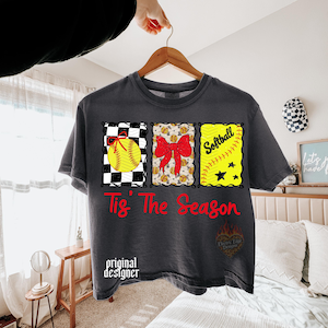 May include: A charcoal grey t-shirt featuring a graphic with three rectangular panels containing a yellow softball with a red bow, a red bow over a softball pattern, and the word Softball with a ball and stars. The text Tis' The Season is in red.