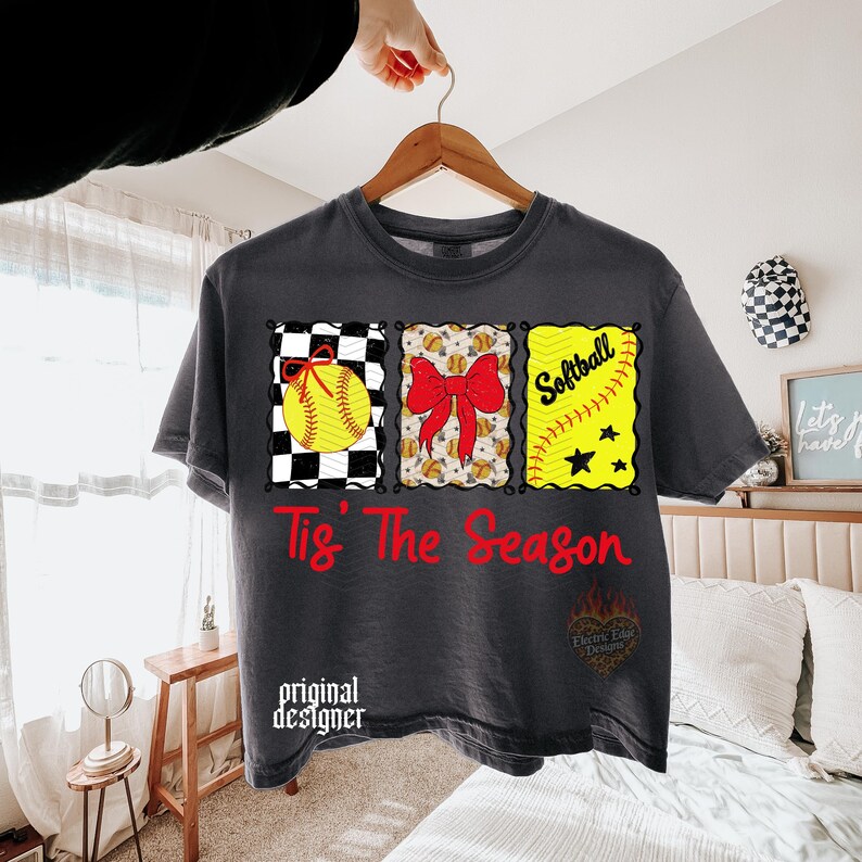 May include: A charcoal grey t-shirt featuring a graphic with three rectangular panels containing a yellow softball with a red bow, a red bow over a softball pattern, and the word Softball with a ball and stars. The text Tis' The Season is in red.