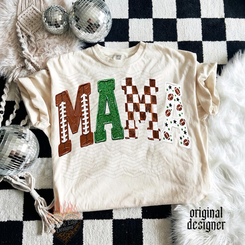 May include: A cream-colored t-shirt featuring the word MAMA in large letters. The M is brown with white football laces, the A is green glitter, the second M is a brown and white checkerboard pattern, and the final A has footballs and stars.
