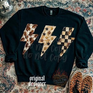 May include: Black sweatshirt featuring three lightning bolt designs with various patterns: a brown and white spotted pattern, gold stars, and a black and white checkered pattern. The text "original designer" is printed at the bottom.
