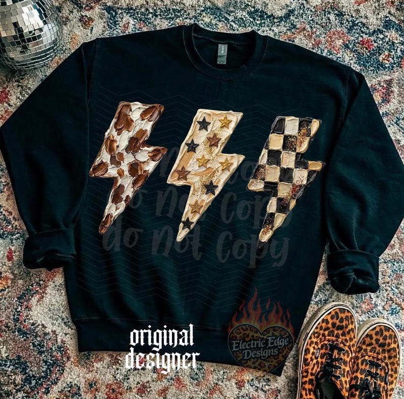 May include: Black sweatshirt featuring three lightning bolt designs with various patterns: a brown and white spotted pattern, gold stars, and a black and white checkered pattern. The text "original designer" is printed at the bottom.
