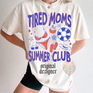 May include: Cream-colored t-shirt with the text "TIRED MOMS SUMMER CLUB" in blue and red. The design includes summer-themed graphics like a bikini, sunglasses, and a beach ball. The words "original designer" are also printed on the shirt.