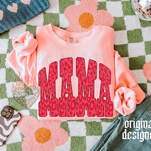May include: A peach-colored long-sleeve crewneck sweatshirt featuring the word MAMA in a bold, red and pink leopard print pattern across the chest. The garment has a ribbed neckline, cuffs, and hem, with a visible brand tag at the inner neck.