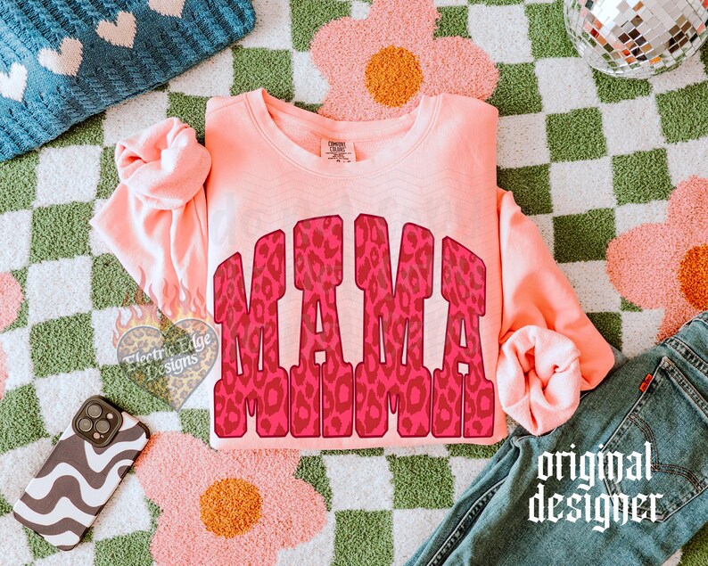 May include: A peach-colored long-sleeve crewneck sweatshirt featuring the word MAMA in a bold, red and pink leopard print pattern across the chest. The garment has a ribbed neckline, cuffs, and hem, with a visible brand tag at the inner neck.