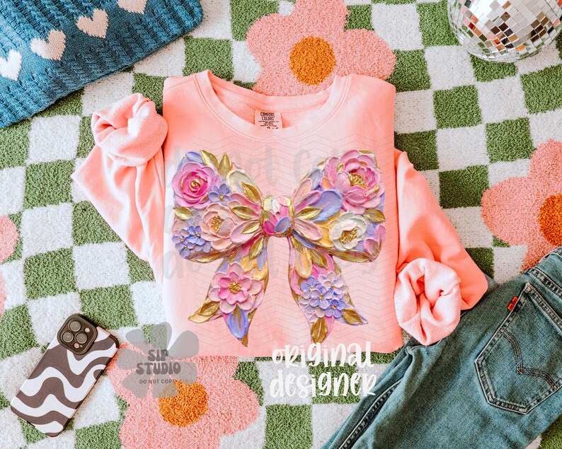 Puede incluir: Una sudadera de color coral con un gran dise&ntilde;o de lazo floral con detalles en rosa, morado y dorado. La frase "original designer" est&aacute; impresa en la prenda. La imagen incluye vaqueros, una funda de tel&eacute;fono y una alfombra estampada.