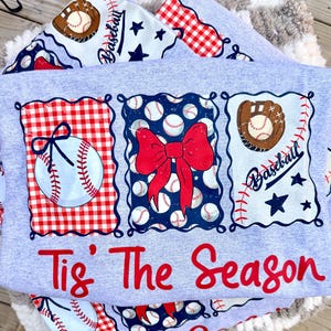 May include: A light gray t-shirt featuring three rectangular panels with baseball-themed graphics. The panels display a baseball with a bow, a red bow over baseballs, and a baseball glove with the word Baseball and stars. Text reads Tis' The Season.