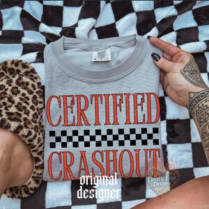 May include: A gray crew neck sweatshirt with the words "CERTIFIED CRASHOUT" in red, with a black and white checkered pattern. The words "original designer" are below the graphic. The sweatshirt is on a black and white checkered blanket.