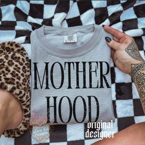 May include: A light gray crewneck t-shirt featuring the black text MOTHER HOOD in a distressed serif font. The shirt has a subtle chevron texture pattern and a white interior neck label. The design is centered on the front of the garment.