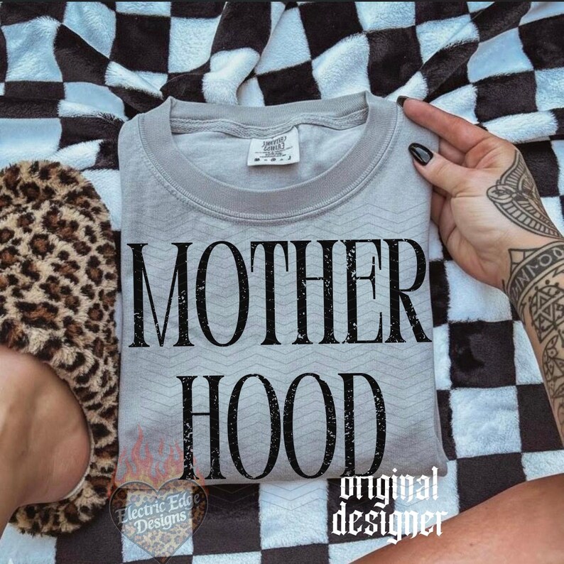 May include: A light gray crewneck t-shirt featuring the black text MOTHER HOOD in a distressed serif font. The shirt has a subtle chevron texture pattern and a white interior neck label. The design is centered on the front of the garment.