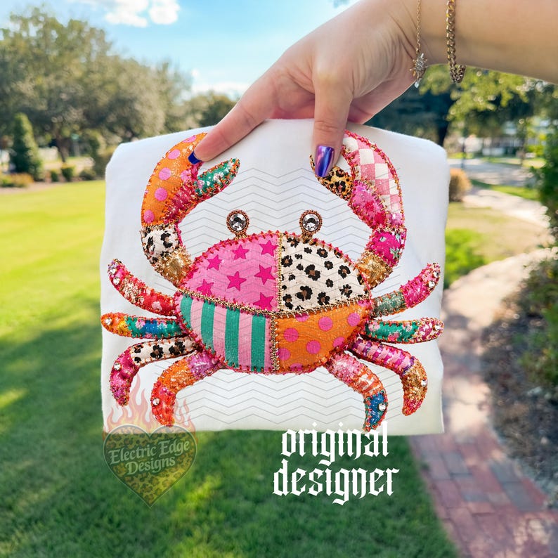 May include: A white t-shirt featuring a colorful crab design. The crab is made of patchwork fabric with various patterns, including stars, polka dots, and stripes. The text "original designer" is printed on the shirt.