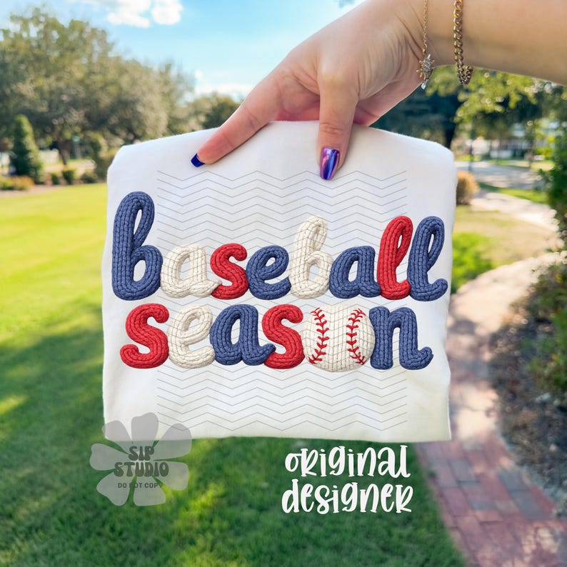 May include: White t-shirt with the words "Baseball Season" in a 3D embroidered style. The letters are in red, white, and blue, with a baseball graphic replacing the letter "O". The shirt is held up by a person.