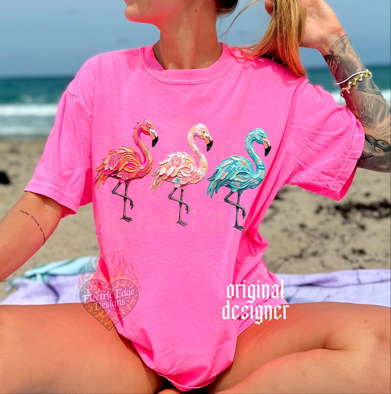 May include: Bright pink t-shirt featuring three flamingo designs in shades of orange, pink, and teal. The shirt has the text "Electric Edge Designs" and "original designer" printed on it. The shirt is made of a soft material.