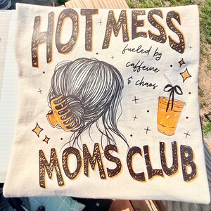 May include: Cream-colored pillow with the words "HOTMESS MOMS CLUB" in brown and black lettering. The design includes an illustration of a woman's hair, a coffee cup, and the phrase "fueled by caffeine & chaos."