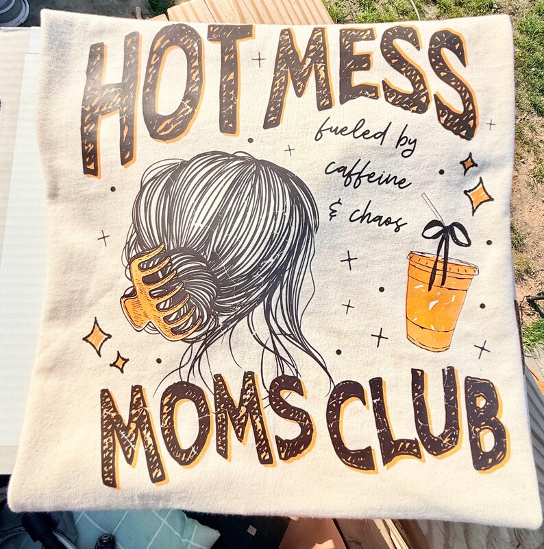 May include: Cream-colored pillow with the words "HOTMESS MOMS CLUB" in brown and black lettering. The design includes an illustration of a woman's hair, a coffee cup, and the phrase "fueled by caffeine & chaos."