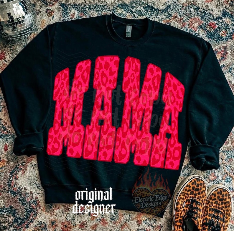 May include: A black long-sleeve crewneck sweatshirt featuring the word MAMA in large, bold, arched block letters. The letters have a vibrant pink and red leopard print pattern. The text original designer and a heart-shaped flame logo are visible.