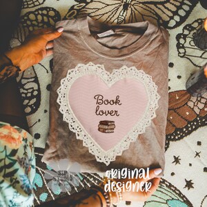 May include: A brown t-shirt with a pink heart-shaped design featuring the words "Book lover" and a stack of books. The heart is outlined with white lace. The words "original designer" are also visible.