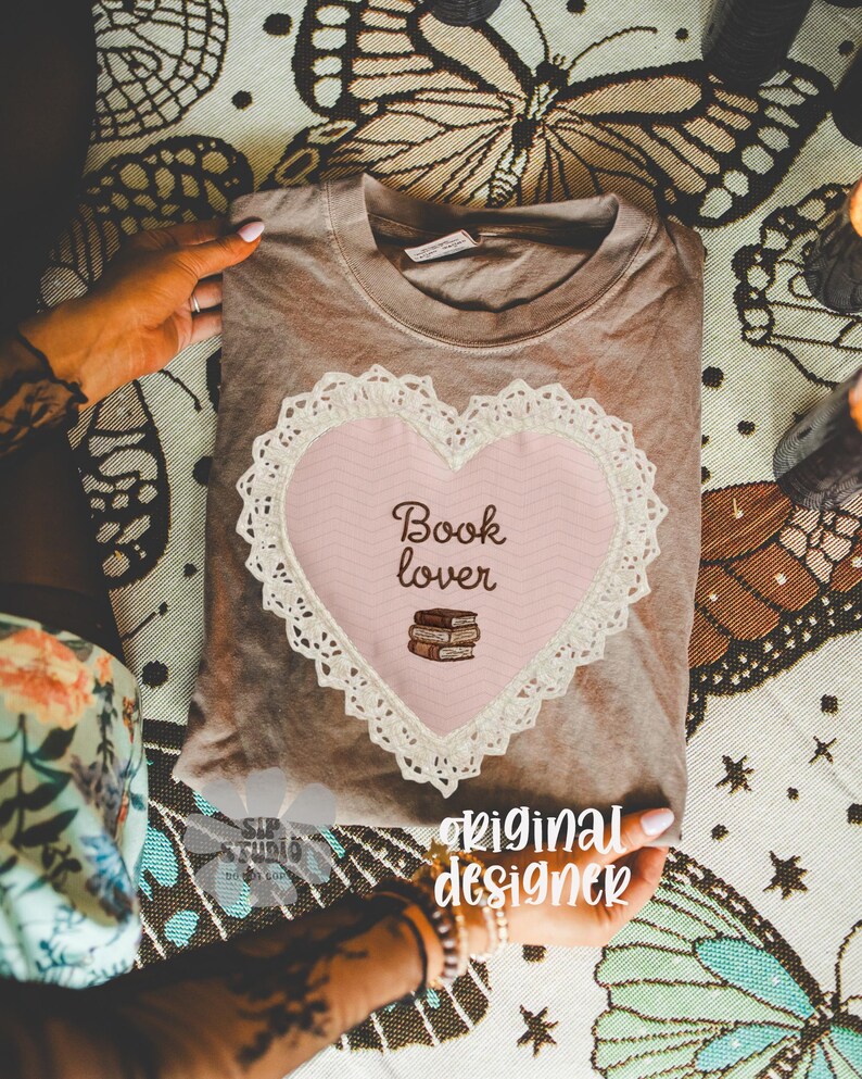May include: A brown t-shirt with a pink heart-shaped design featuring the words "Book lover" and a stack of books. The heart is outlined with white lace. The words "original designer" are also visible.