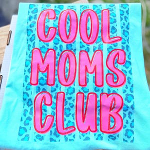 Peut inclure: T-shirt turquoise avec l'inscription "COOL MOMS CLUB" en grandes lettres roses bord&eacute;es de rouge. Le fond pr&eacute;sente un motif l&eacute;opard bleu et des rayures verticales.