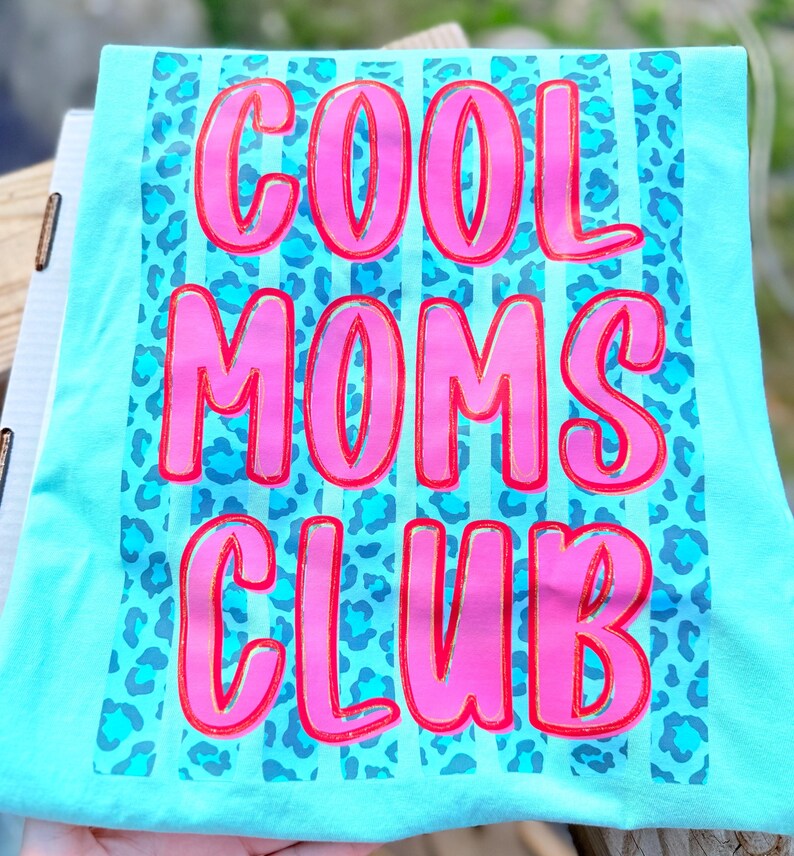 Peut inclure: T-shirt turquoise avec l'inscription "COOL MOMS CLUB" en grandes lettres roses bord&eacute;es de rouge. Le fond pr&eacute;sente un motif l&eacute;opard bleu et des rayures verticales.