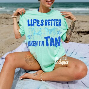 May include: A light green t-shirt with the text "LIFE'S BETTER WHEN I'M TAN" in blue. The shirt features a design of hanging swimsuits and stars. The image is taken on a beach.