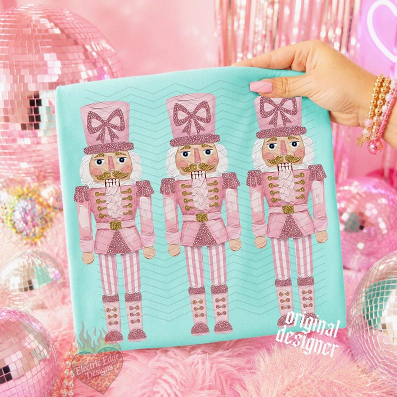May include: A light turquoise t-shirt featuring a repeating graphic of three pink nutcracker figures. Each nutcracker wears a tall hat with a glittery pink bow, a gold mustache, and a pink uniform with gold accents. The fabric has a subtle chevron pattern.