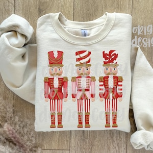 May include: Cream-colored sweatshirt featuring three nutcracker soldier designs. Each nutcracker is dressed in a red and white uniform with a matching hat. The design is centered on the front of the sweatshirt, with the sleeves rolled up.