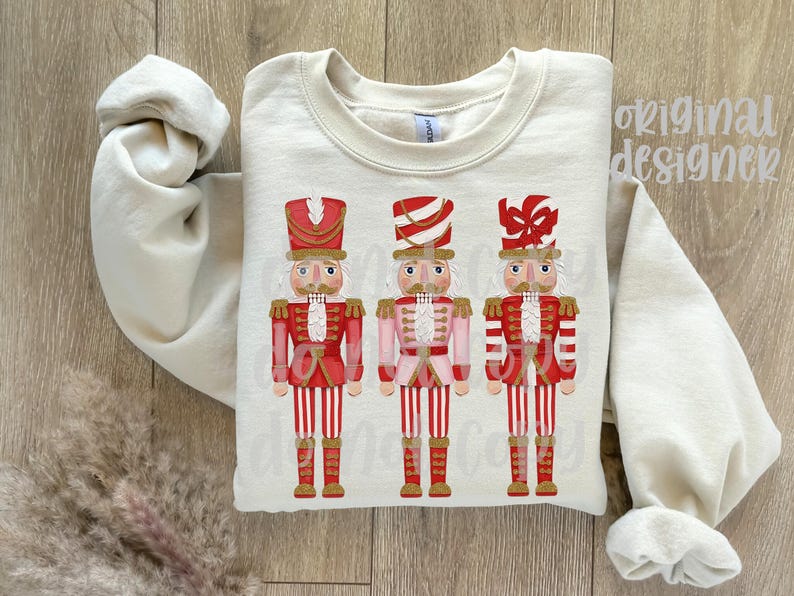 May include: Cream-colored sweatshirt featuring three nutcracker soldier designs. Each nutcracker is dressed in a red and white uniform with a matching hat. The design is centered on the front of the sweatshirt, with the sleeves rolled up.