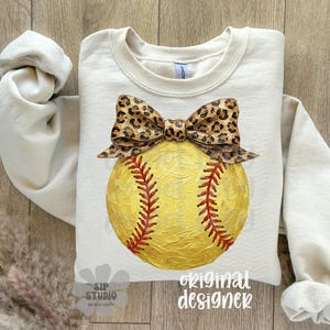 Op de afbeelding: Cr&egrave;mekleurige sweatshirt met een geel softbalontwerp, rode stiksels en een luipaardprint strik. De top met lange mouwen heeft een ronde hals. De woorden "original designer" staan rechtsonder gedrukt. Geschikt voor sportfans.