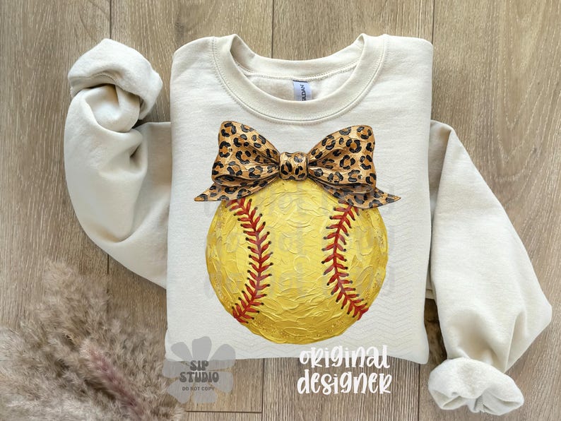Op de afbeelding: Cr&egrave;mekleurige sweatshirt met een geel softbalontwerp, rode stiksels en een luipaardprint strik. De top met lange mouwen heeft een ronde hals. De woorden "original designer" staan rechtsonder gedrukt. Geschikt voor sportfans.