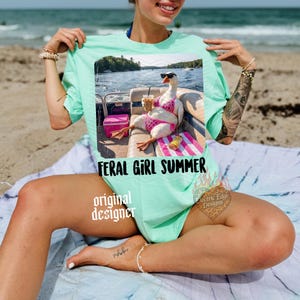 K&ouml;nnte beinhalten: Mintgr&uuml;nes T-Shirt mit einem Aufdruck einer Gans mit Sonnenbrille und pinkem Bikini auf einem Boot. Der Text "FERAL GIRL SUMMER" steht darunter. Das T-Shirt hat eine lockere Passform und ist f&uuml;r Freizeitkleidung geeignet.